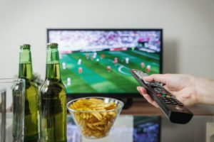 Two beers, bowl of chips, hand holding remote to TV playing sporting event
