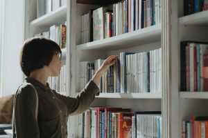 Woman stands near white bookshelf full of books and selects a book.