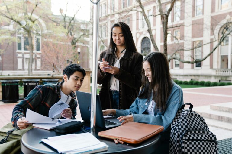 A group of students sits at a table outside exchanging notes.