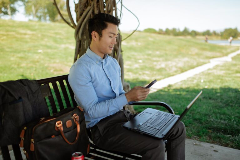 Man in Blue Long Sleeve Shirt Using Cellphone with computer in lap on park bench
