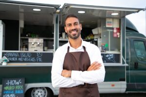 Cheerful male food truck waiter standing with crossed arms in front of truck.