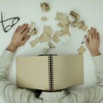A Man Resting on a Table with a notebook on his head, pen in hand and shredded paper in front of him