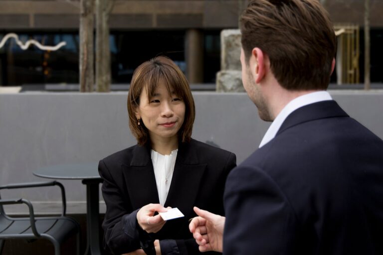 Smiling Businessman and Businesswoman Exchanging a Business Card at a Table in Front of an Office Building