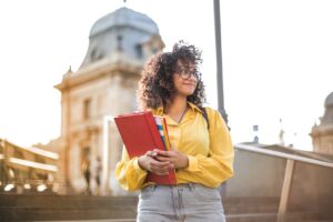 Woman In Yellow Jacket Holding Books in front of college campus building