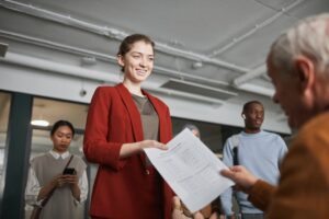 A Woman in Red Blazer handing White Paper to man at desk