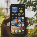 Close-up of a smartphone held outdoors in an autumn setting, showcasing apps and widgets.
