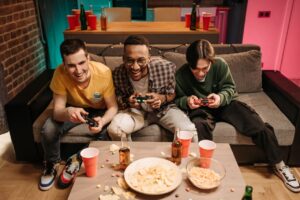 Three men playing video games together on a sofa with snacks and drinks around.