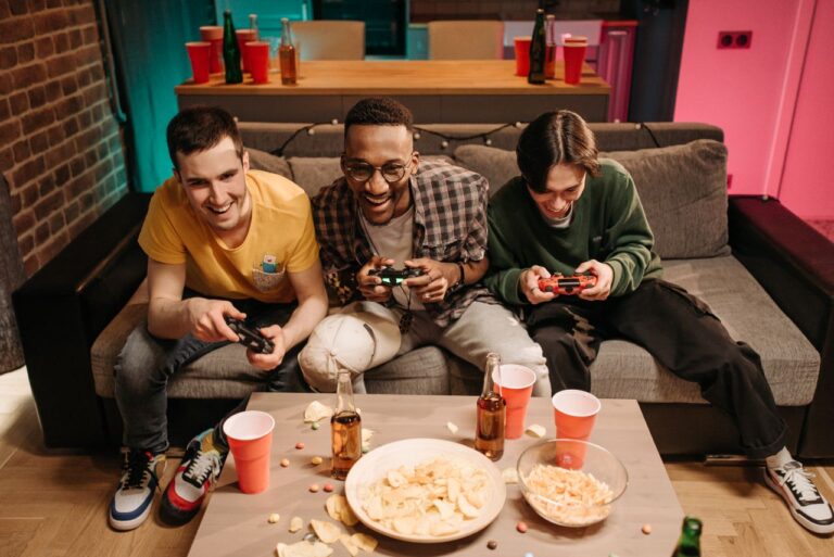 Three men playing video games together on a sofa with snacks and drinks around.