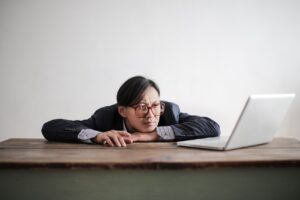 Asian businessman in glasses, resting head on desk, looking at laptop in office setting.